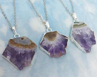 Puede incluir: Cuatro collares de plata con colgantes de cristal de amatista. Los cristales de amatista son en bruto y tienen un color morado y blanco con inclusiones marrones. Los collares están sobre un fondo azul claro.