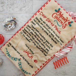 May include: A cream-colored pet bed with a red border and a letter to Santa Paws. The letter includes the text "Merry Christmas" and a request for treats and toys. A red and white striped ribbon is attached.