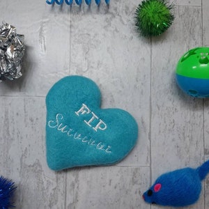 May include: A blue heart-shaped cat toy with the text "FIP Survivor" embroidered on it. The toy is surrounded by other cat toys, including a silver ball, a green ball with holes, a blue and green ball, and a blue fuzzy mouse.