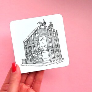 May include: Black and white line drawing of a building with the text "The Fat Cat" above the entrance. The drawing is on a white coaster.