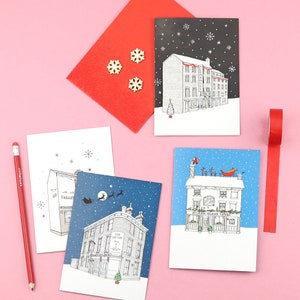 May include: A collection of Christmas cards featuring black and white illustrations of buildings in a snowy landscape. The cards are accompanied by a red envelope, wooden snowflakes, a red pencil, and red tape, all set against a pink background.