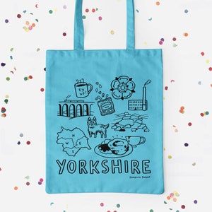 Puede incluir: Una bolsa de tela azul claro con un diseño negro que representa lugares emblemáticos de Yorkshire, como un pudín de Yorkshire, una taza de té, un perro y la rosa de Yorkshire. El texto "Yorkshire" está impreso en negro en la bolsa.