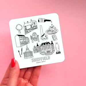May include: A white coaster with black line drawings of Sheffield landmarks, including a building with a clock tower, a park, a pub, and a bottle of wine. The text "SHEFFIELD" is written in black at the bottom of the coaster.