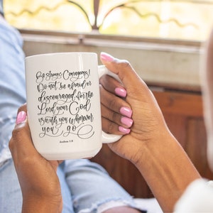 May include: White ceramic mug with the text "Be strong and courageous! Do not be afraid or discouraged, for the Lord your God is with you wherever you go." in black script. The mug has a large handle and is held by a person.