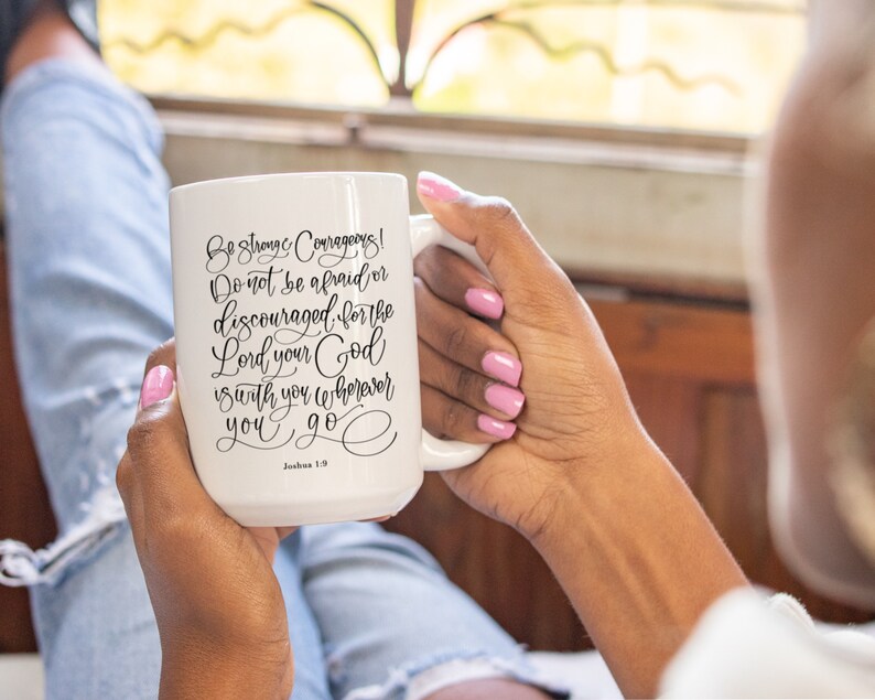 May include: White ceramic mug with the text "Be strong and courageous! Do not be afraid or discouraged, for the Lord your God is with you wherever you go." in black script. The mug has a large handle and is held by a person.