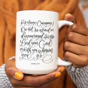 May include: White ceramic mug with a large handle, featuring the scriptural quote "Be Strong & Courageous! Do not be afraid or discouraged for the Lord your God is with you wherever you go" from Joshua 1:9 in black script.