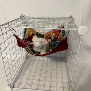 Op de afbeelding: Een witte draadkooi met een rode en bedrukte hangmat erin. Een klein, wit knuffeldier rust in de hangmat. De hangmat is gemaakt van stof met een Harry Potter boekomslagontwerp.
