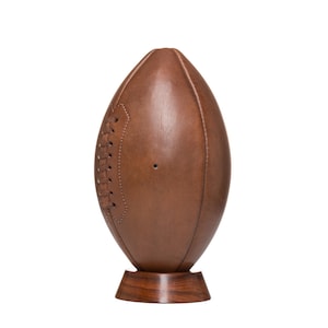 May include: A brown leather football on a wooden stand. The football has white stitching and a brown leather texture.