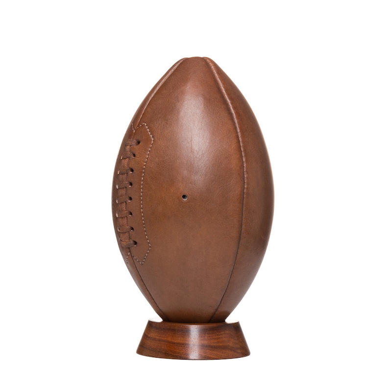 May include: A brown leather football on a wooden stand. The football has white stitching and a brown leather texture.