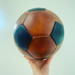 K&ouml;nnte beinhalten: Ein Fu&szlig;ball im Vintage-Stil mit braunen und marineblauen Paneelen. Der Ball ist aus Leder gefertigt und hat ein klassisches Design. Er wird in einer Hand vor einem schlichten Hintergrund gehalten.