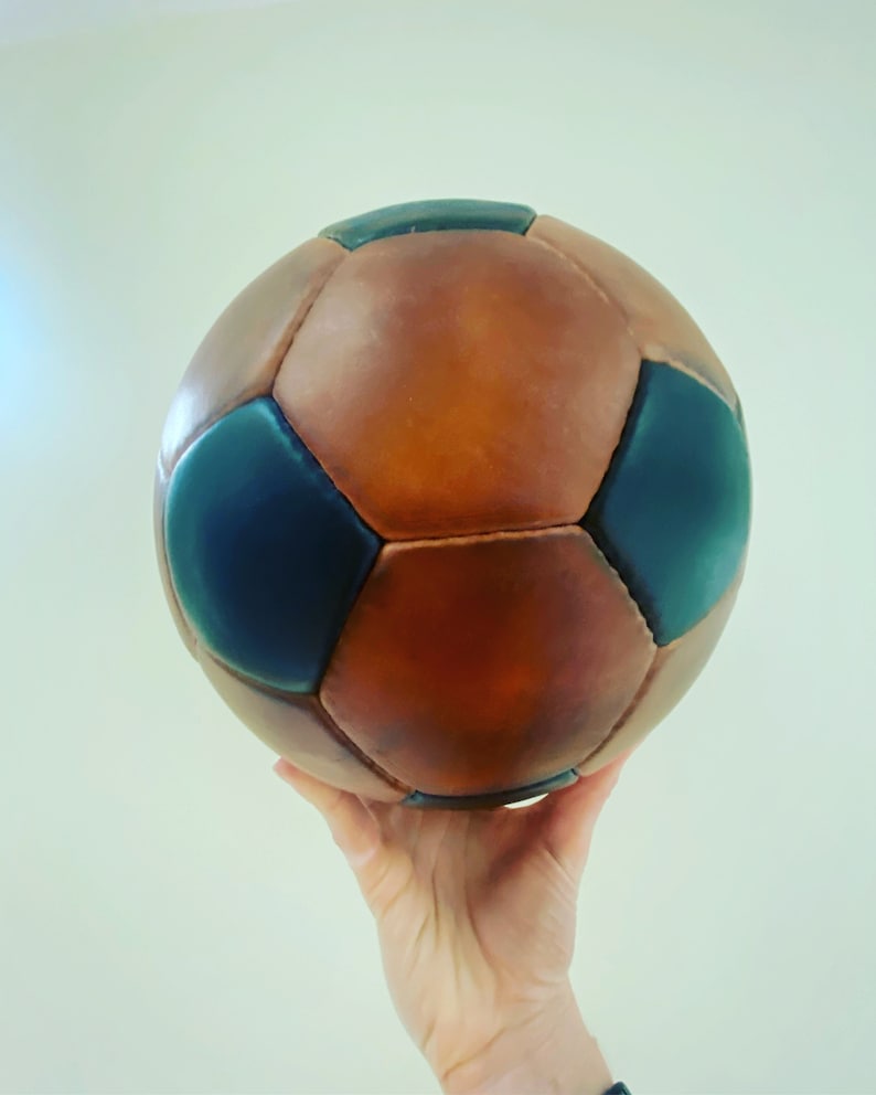 K&ouml;nnte beinhalten: Ein Fu&szlig;ball im Vintage-Stil mit braunen und marineblauen Paneelen. Der Ball ist aus Leder gefertigt und hat ein klassisches Design. Er wird in einer Hand vor einem schlichten Hintergrund gehalten.