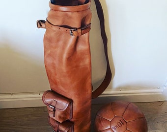 May include: A brown leather golf bag with a strap and two pockets. The bag is standing upright with a golf club sticking out of the top. There are two brown leather medicine balls in the foreground.