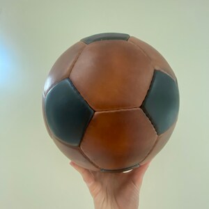 K&ouml;nnte beinhalten: Ein braun-schwarzer Fu&szlig;ball mit klassischem Design. Der Ball ist aus Leder gefertigt und hat ein traditionelles F&uuml;nfeck- und Sechseckmuster. Der Ball wird in der Luft gehalten.