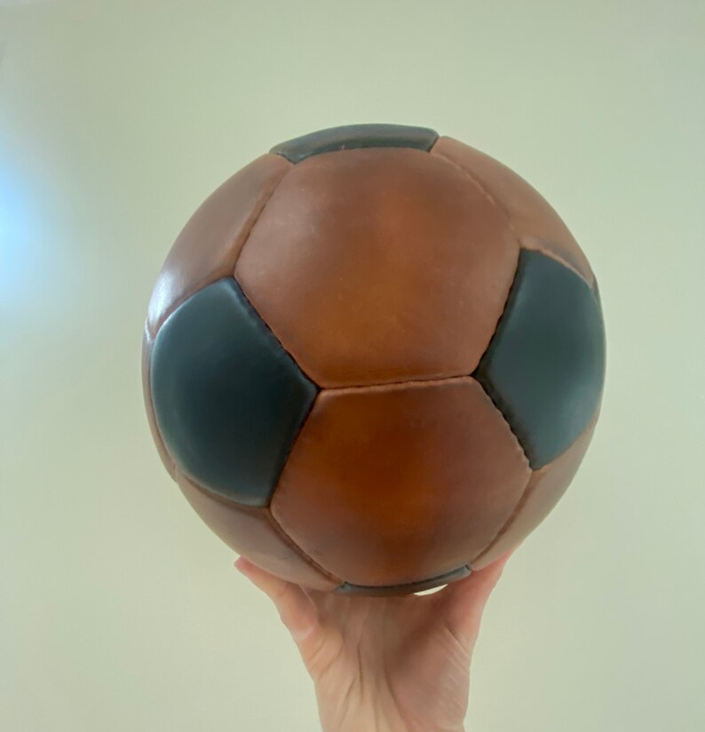 K&ouml;nnte beinhalten: Ein braun-schwarzer Fu&szlig;ball mit klassischem Design. Der Ball ist aus Leder gefertigt und hat ein traditionelles F&uuml;nfeck- und Sechseckmuster. Der Ball wird in der Luft gehalten.