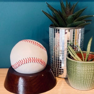 Pode incluir: Uma bola de beisebol branca com costuras vermelhas repousa sobre uma tigela de madeira escura. Uma planta verde num vaso texturizado e um vaso espelhado estão no fundo.