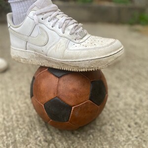 K&ouml;nnte beinhalten: Ein wei&szlig;er Ledersneaker liegt auf einem braunen und schwarzen Fu&szlig;ball. Der Schuh hat eine wei&szlig;e Sohle und wei&szlig;e Schn&uuml;rsenkel. Der Fu&szlig;ball hat ein klassisches F&uuml;nfeck- und Sechseck-Design.