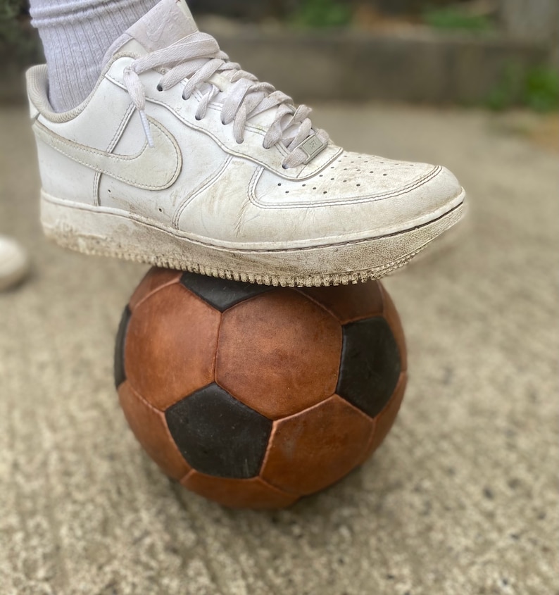 K&ouml;nnte beinhalten: Ein wei&szlig;er Ledersneaker liegt auf einem braunen und schwarzen Fu&szlig;ball. Der Schuh hat eine wei&szlig;e Sohle und wei&szlig;e Schn&uuml;rsenkel. Der Fu&szlig;ball hat ein klassisches F&uuml;nfeck- und Sechseck-Design.
