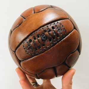 May include: A vintage brown leather soccer ball with a stitched panel design. The ball has a black rubber valve.