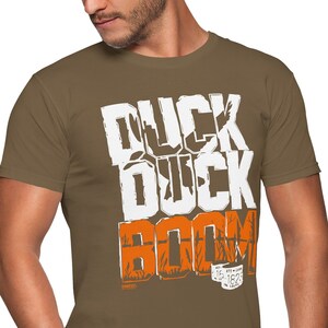 Op de afbeelding: Een bruin T-shirt met de tekst "Duck Duck Boom" bedrukt in wit en oranje. De tekst is gestyled met een eenden silhouet en een jachtsc&egrave;ne.