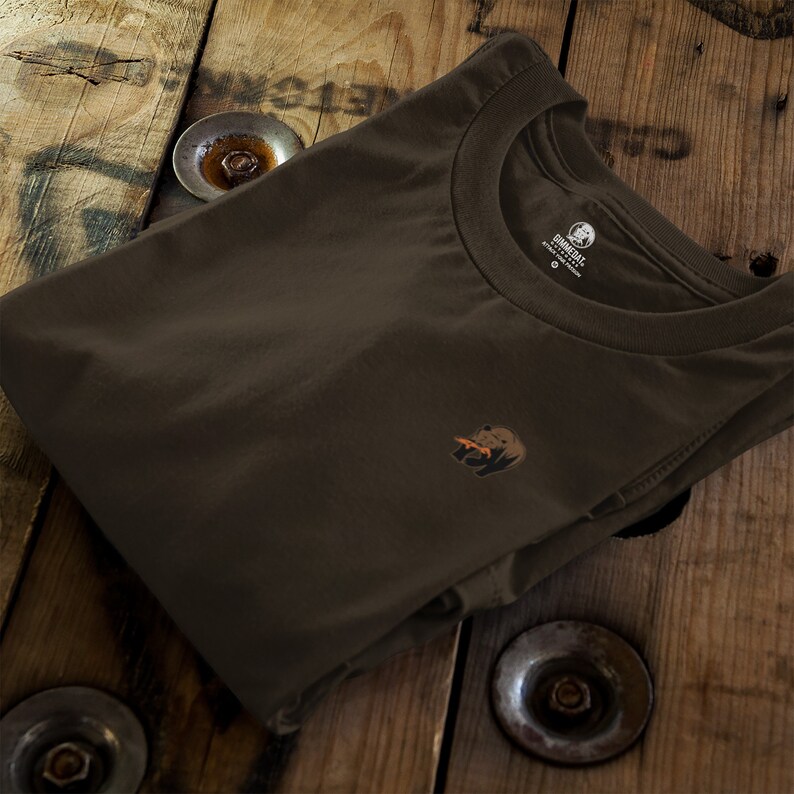 May include: A brown t-shirt with a small graphic of a bear eating a donut. The shirt is folded and laid on a wooden surface.