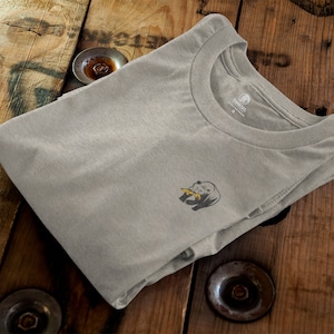 May include: A gray t-shirt with a small graphic of a rhinoceros on the left chest. The rhinoceros is gray with a yellow horn and is eating a leaf. The t-shirt has a crew neck and is folded in half.