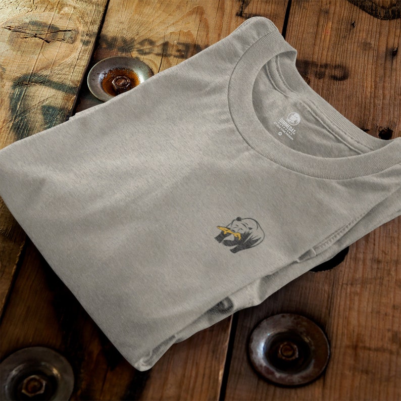 May include: A gray t-shirt with a small graphic of a rhinoceros on the left chest. The rhinoceros is gray with a yellow horn and is eating a leaf. The t-shirt has a crew neck and is folded in half.