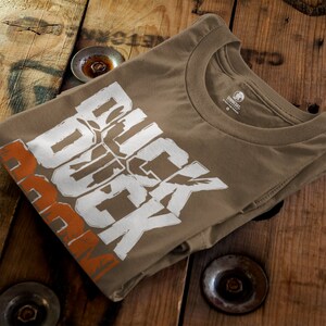 Op de afbeelding: Een bruin t-shirt met een witte afbeelding waarop in een vetgedrukte, gestileerde lettertype "Duck Duck Boom" staat. De afbeelding bevat ook een gestileerd vogelontwerp.