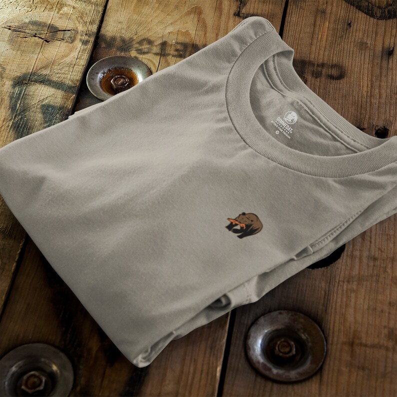 May include: A light gray t-shirt with a small brown bear graphic wearing a slice of orange. The shirt has a crew neck and is folded on a wooden surface.