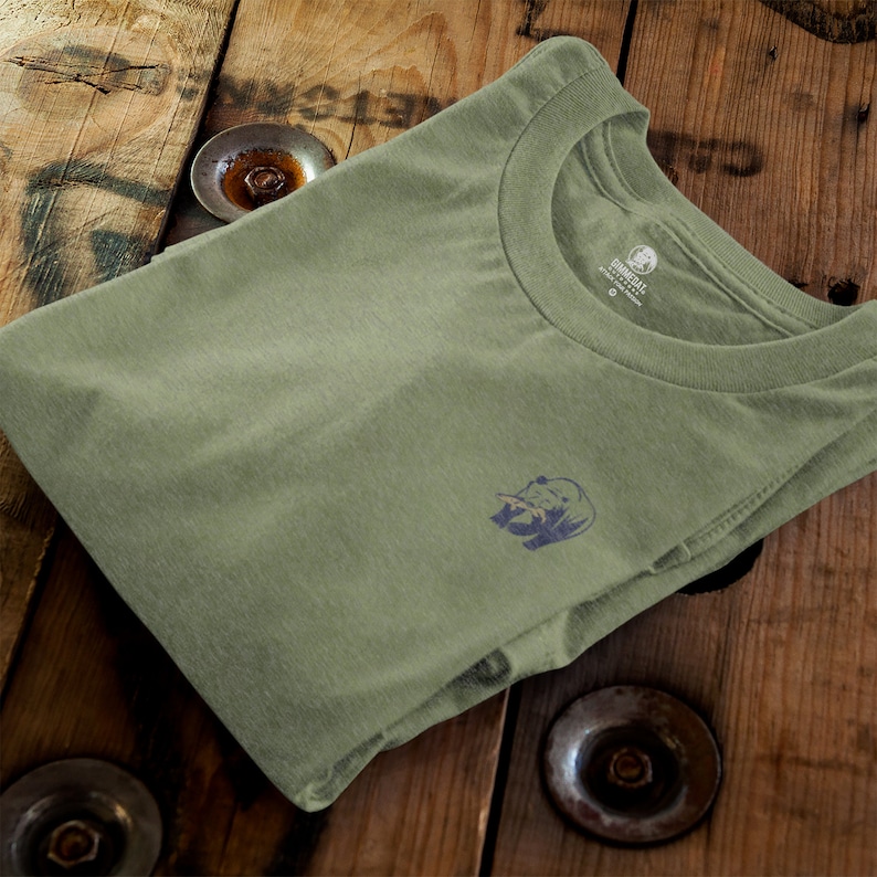 K&ouml;nnte beinhalten: Ein gefaltetes gr&uuml;nes T-Shirt mit einem kleinen blauen B&auml;ren-Motiv auf der linken Seite. Das Shirt hat einen Rundhalsausschnitt und ist aus weichem, bequemem Stoff gefertigt. Der Markenname "DOVERBELL" ist auf dem Etikett sichtbar.
