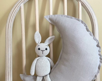 May include: A crocheted bunny rabbit sits on a light brown crescent moon shaped pillow. The pillow is on a wooden chair with a light yellow wall in the background.