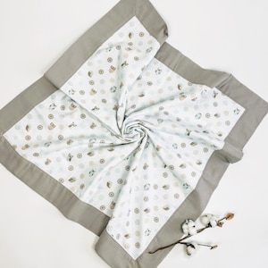 May include: A white baby blanket with a gray border and a pattern of owls and circles. The blanket is folded and twisted, and a sprig of cotton is in the lower right corner. The text "CottonByAlena" is in the lower right corner.
