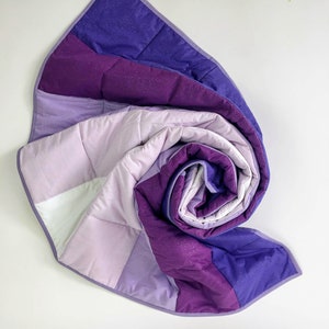 May include: A purple and white quilted blanket with a sparkly texture is rolled up in a spiral shape. The blanket is made of soft, comfortable fabric and has a modern design.