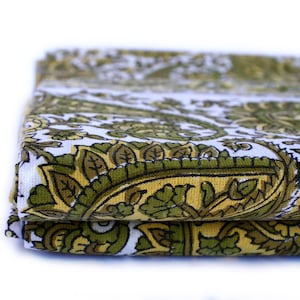 May include: Folded white fabric with a repeating paisley pattern in shades of green and yellow. The fabric has a detailed floral design with black outlines, creating a layered effect.