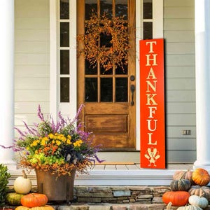May include: A red wooden sign with the word "THANKFUL" in gold lettering. A gold turkey illustration is at the bottom of the sign. The sign is leaning against a white door with a fall wreath. Pumpkins are on the steps in front of the door.