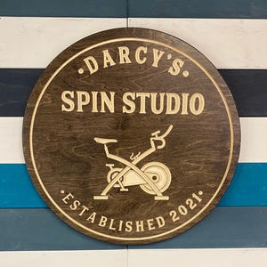 May include: A round wooden sign with the text "Darcy's Spin Studio Established 2021." The sign features a stylized image of a stationary bike.