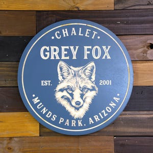 May include: A blue round wooden sign with the text "•CHALET. GREY FOX EST. 2001 •MUNDS PARK, ARIZONA." in white lettering. A white silhouette of a fox's head is in the center of the sign.