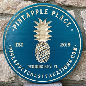 May include: A teal blue round wooden sign with the text "Pineapple Place" in white lettering. The sign also features a carved pineapple in the center, the text "Est. 2019", and the text "Perdido Key, FL Pineapple Coast Vacations.com".