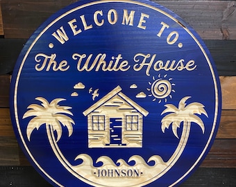 May include: A round wooden sign with a blue background and a white inscription that reads "Welcome to The White House - Johnson". The sign features a stylised image of a beach house with palm trees and waves.