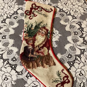 May include: A Christmas stocking with a festive design. The stocking features a detailed needlepoint image of Santa Claus and a child, surrounded by greenery. The stocking has a red trim and a loop for hanging.