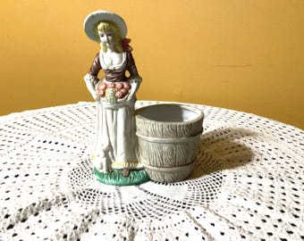 Puede incluir: Un macetero de cerámica blanca con forma de mujer que sostiene una cesta de fruta. El macetero lleva un sombrero blanco y un vestido marrón. El macetero está de pie sobre una base verde. El macetero está sentado sobre un mantel de crochet blanco.