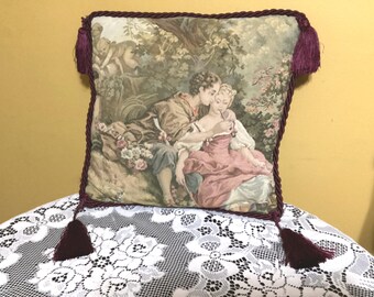Op de afbeelding: Een kussen met een tapijtbekleding, een bordeauxrode rand en kwastjes. De tapisserie toont een stel dat elkaar omarmt in een tuin.