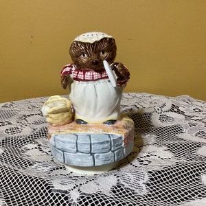 Puede incluir: Una figurita musical de cerámica de un erizo que lleva un vestido a cuadros rojo y blanco y un delantal blanco, sosteniendo un plato blanco. La figurita está de pie sobre una base de piedra con una cesta de flores.