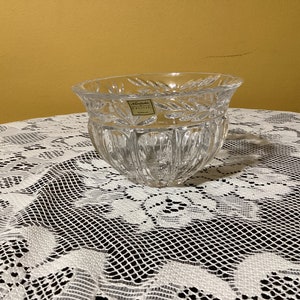 Puede incluir: Un cuenco de vidrio transparente con un patrón decorativo se encuentra sobre un mantel de encaje blanco. El cuenco tiene una etiqueta que dice "Noritake Crystal".