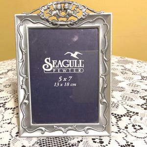 Puede incluir: Un marco de fotos de peltre plateado con un diseño ornamentado que presenta dos querubines en la parte superior. El marco es para una foto de 13 x 18 cm y tiene el texto "Seagull Pewter" en la parte delantera.