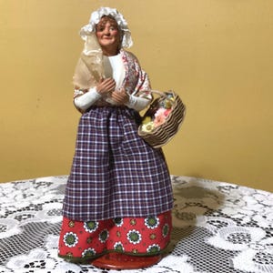 May include: A handmade figurine of a woman wearing a white bonnet, a plaid skirt, and a floral shawl. She is holding a basket filled with flowers.