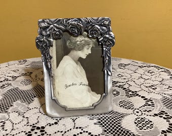 May include: A silver picture frame with a floral design. The frame has a raised design of roses and leaves. The frame is holding a black and white photo of a woman. The text "Pewter Frame" is visible in the photo.