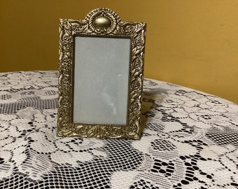 May include: A gold-toned picture frame with an ornate design. The frame has a raised, decorative border and a blank space for a photo.