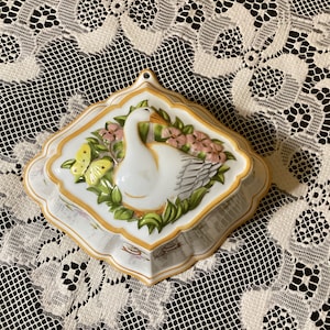 Puede incluir: Un molde de cerámica blanca con forma de ganso, con un borde amarillo y detalles florales. El molde está decorado con una mariposa y flores rosas.