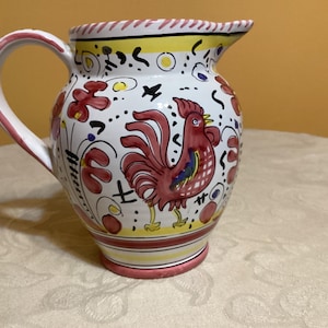 May include: A white ceramic pitcher with a red rooster design. The pitcher has a curved handle and a spout for pouring. The design includes red flowers, yellow accents, and black details. The pitcher is approximately 8 inches tall.