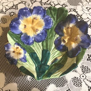 May include: A ceramic dish with a blue, yellow, and green floral design. The dish features three stylized flowers with yellow centers and blue petals.